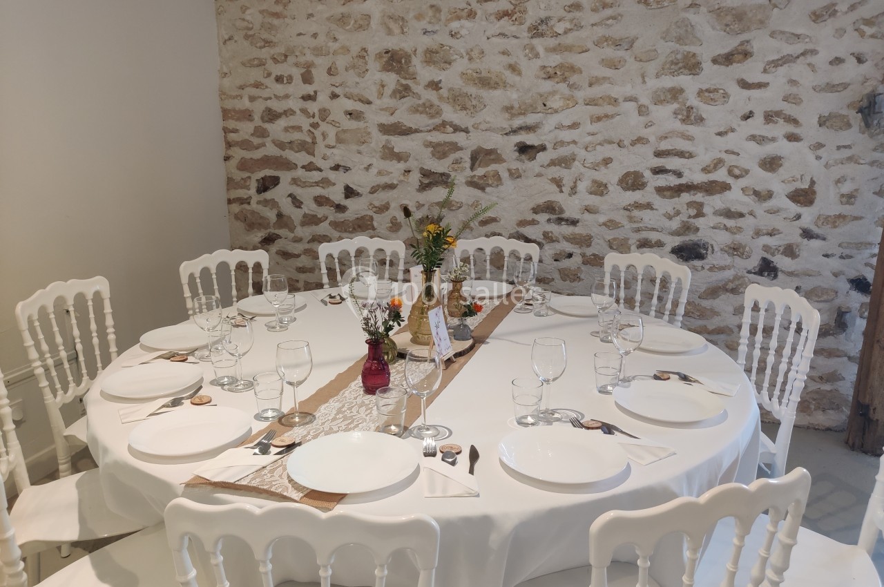 Table ronde dressée avec nappes blanches, vaisselle, couverts et décorations florales, devant un mur en pierre apparente.