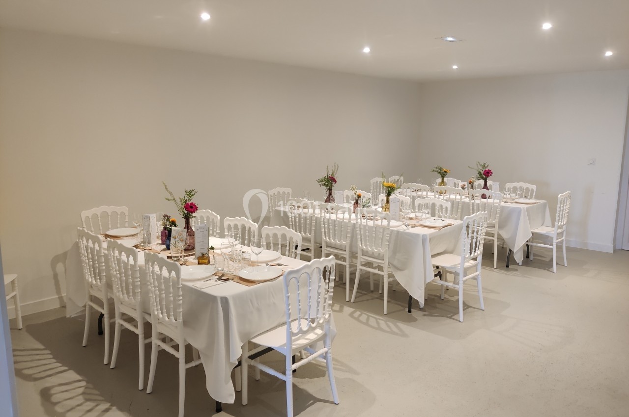 Salle décorée avec des tables rectangulaires dressées, nappes blanches et bouquets de fleurs colorées.