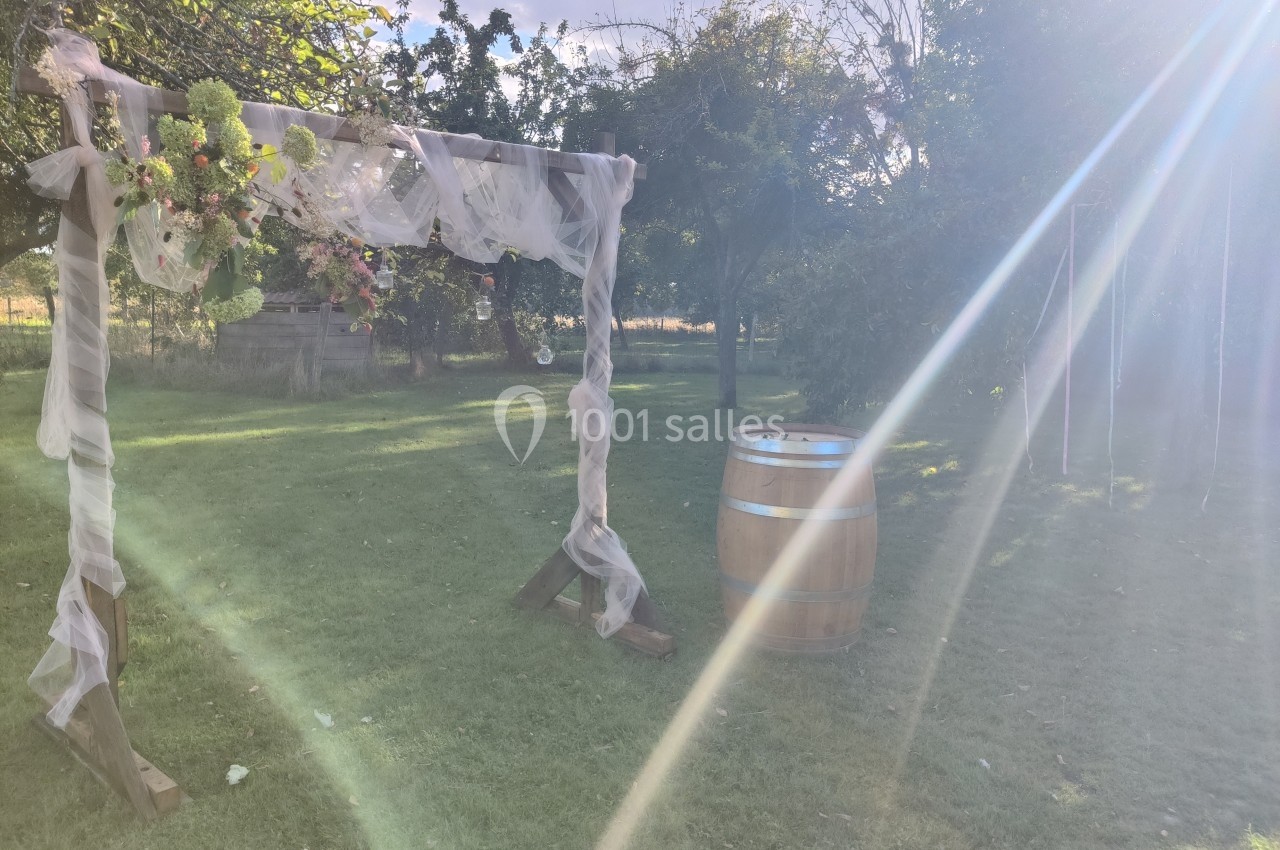 Arche décorée de voilages et fleurs dans un jardin ensoleillé, avec un tonneau en bois à proximité.