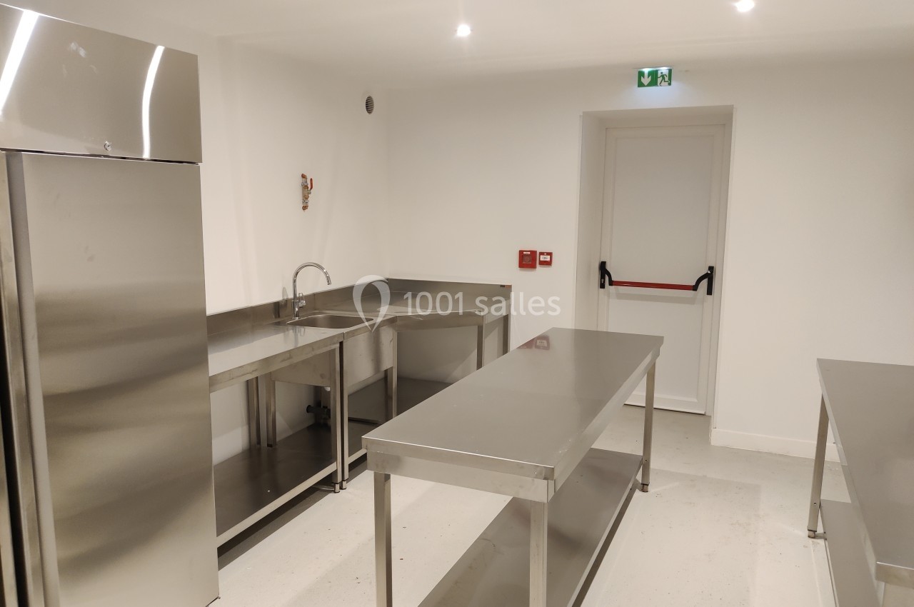 Cuisine professionnelle vide avec plans de travail en inox, évier, réfrigérateur et porte de sortie de secours.