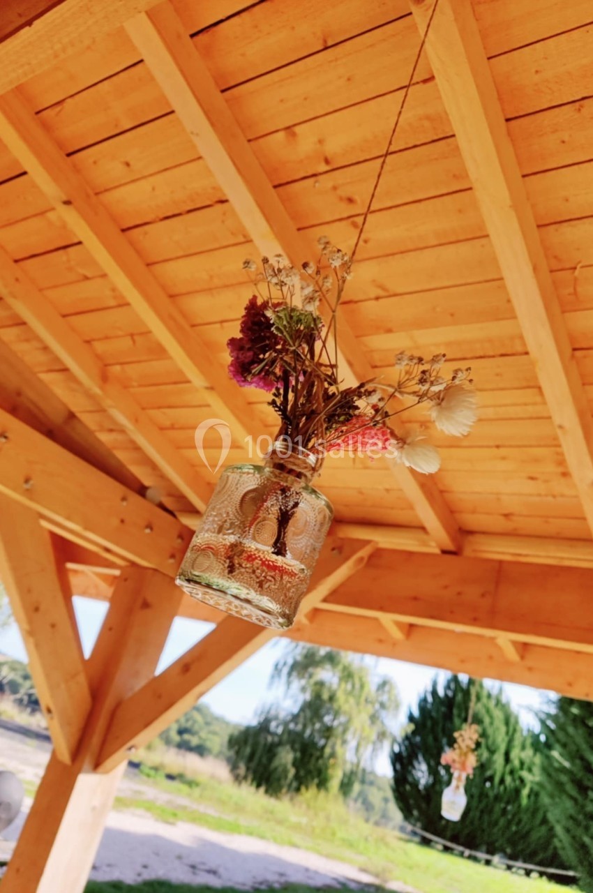 Un petit vase suspendu contenant des fleurs séchées, accroché sous une pergola en bois avec un paysage en arrière-plan.