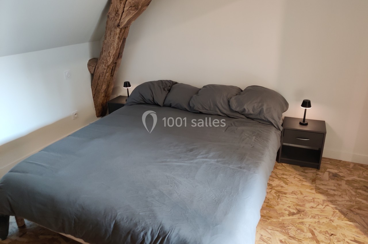 Lit double avec draps gris, encadré par deux tables de chevet noires avec lampes, dans une chambre mansardée.