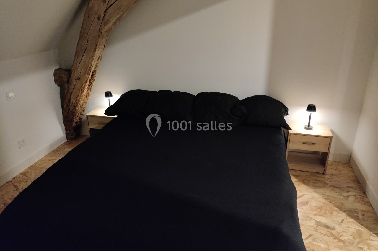 Lit double avec draps noirs, encadré de deux tables de chevet en bois clair avec lampes allumées, dans une chambre mansardée.