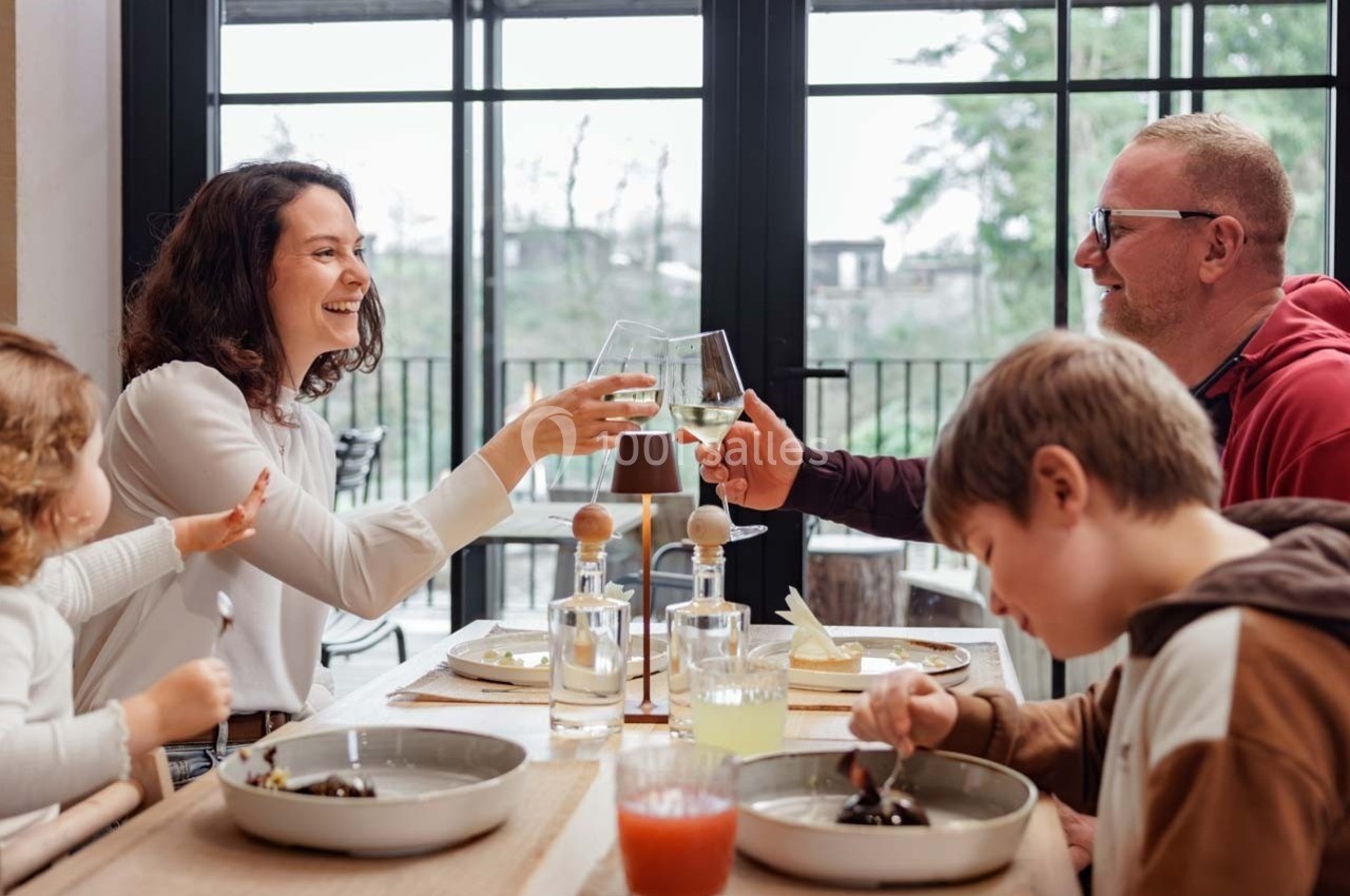 Une famille partage un repas à table, deux adultes trinquent avec des verres de vin blanc dans une salle lumineuse.