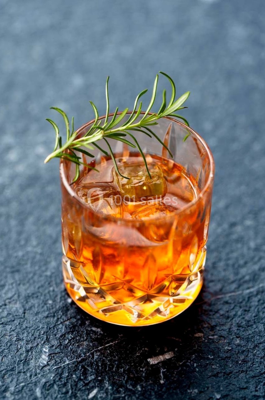 Verre de cocktail ambré avec glaçons, décoré d'une branche de romarin, posé sur une surface sombre.