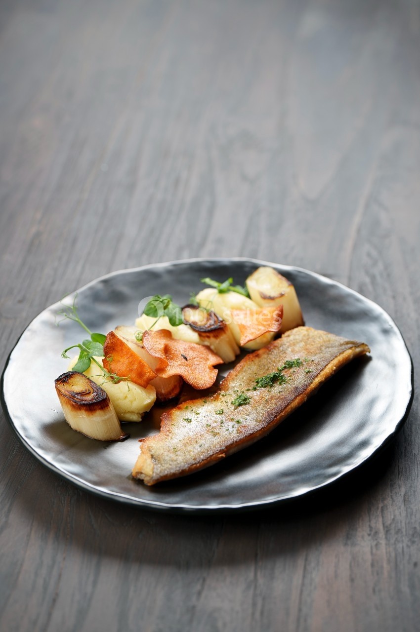 Filet de poisson grillé servi avec des légumes rôtis et des chips de légumes sur une assiette noire.