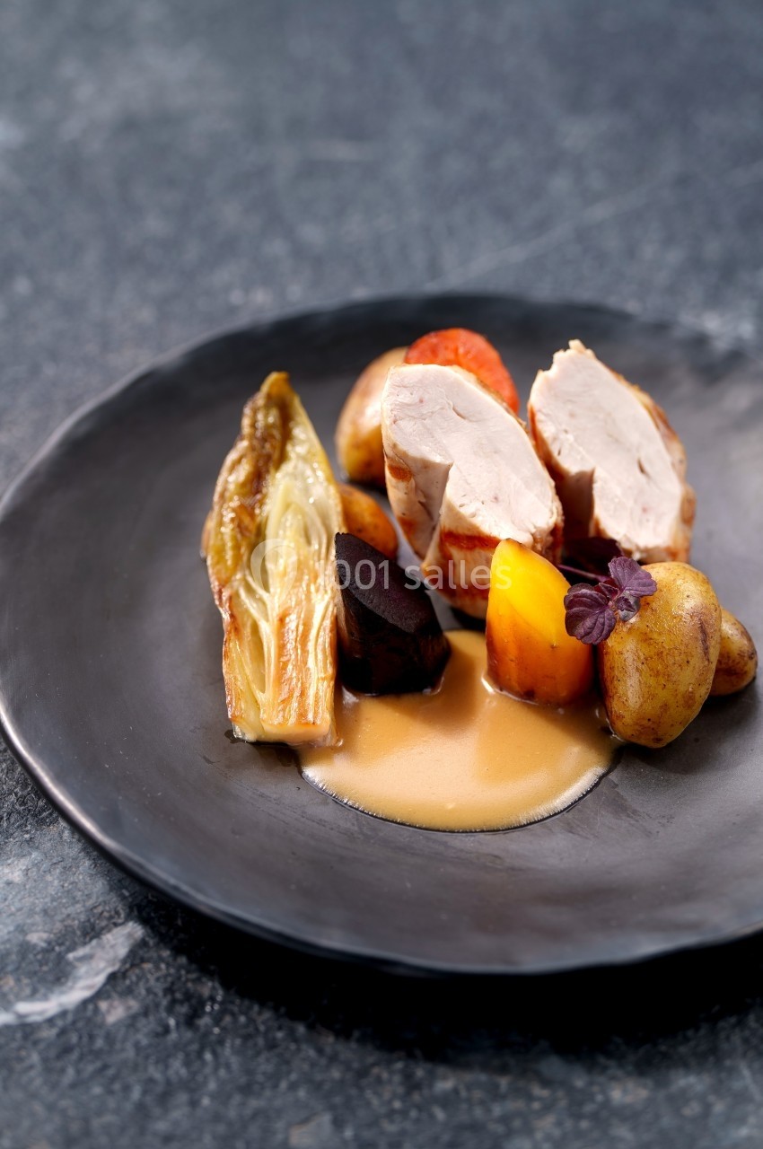 Assiette de volaille tranchée avec légumes racines, pommes de terre, endive braisée et sauce, sur fond sombre.