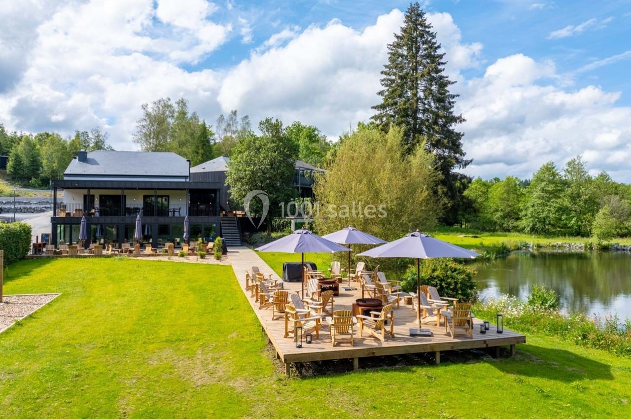 Terrasse en bois avec tables et parasols, bordant un étang entouré de verdure et d'arbres sous un ciel partiellement nuageux.