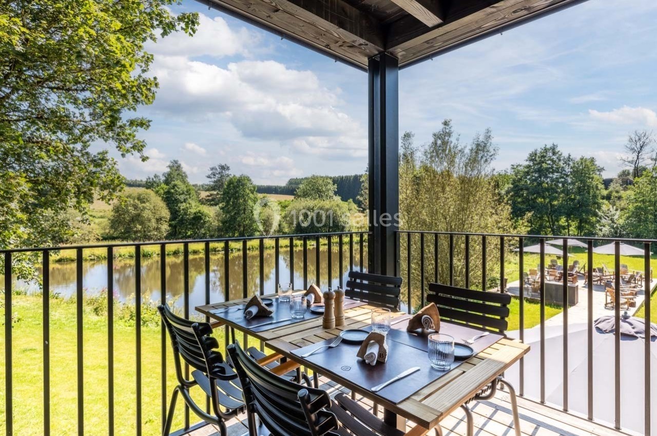 Table en bois dressée sur une terrasse avec vue sur un jardin verdoyant et une rivière sous un ciel partiellement nuageux.