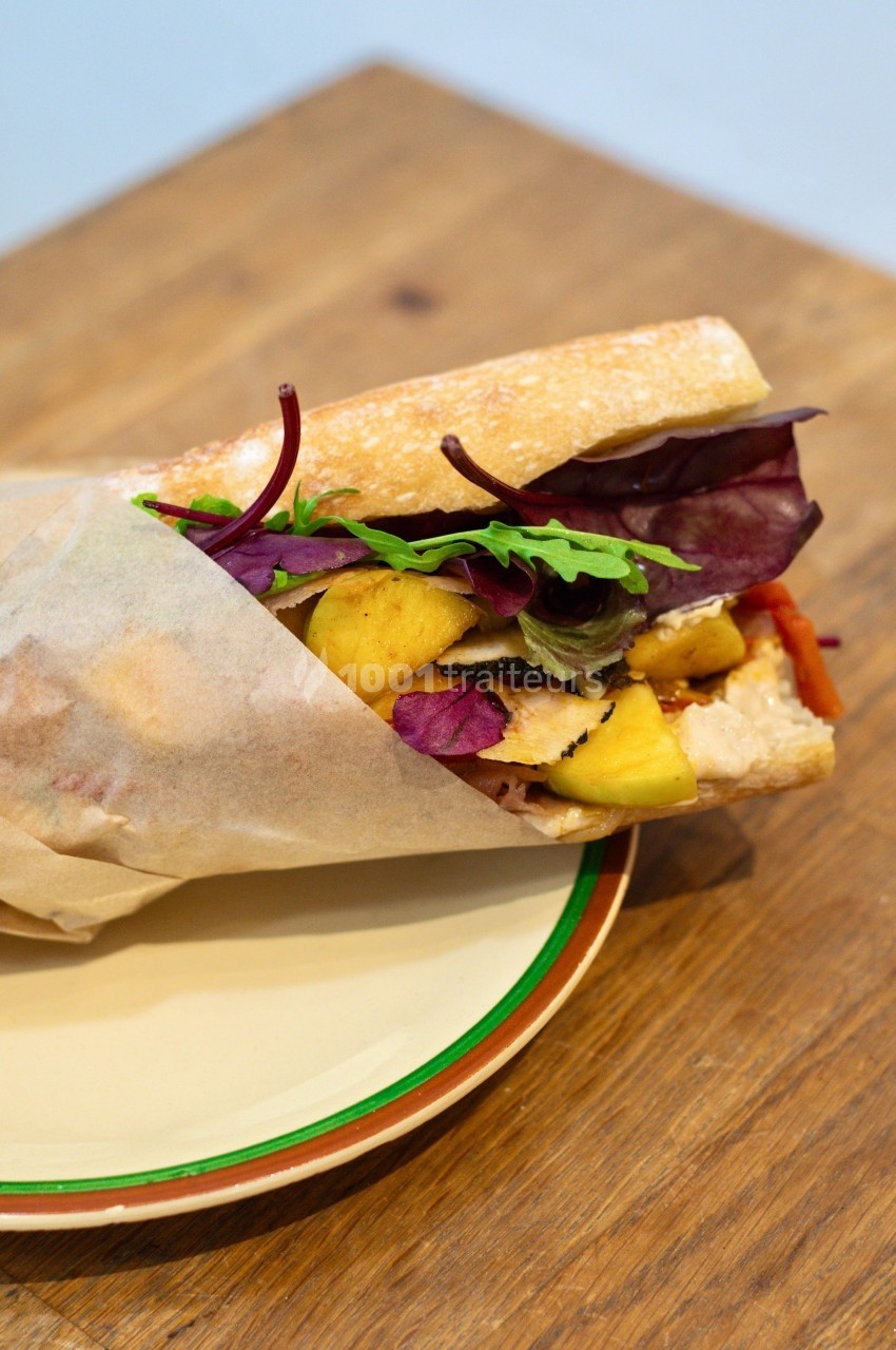 Sandwich garni de poulet, feuilles de salade, morceaux de mangue et légumes, présenté sur une assiette en bois.