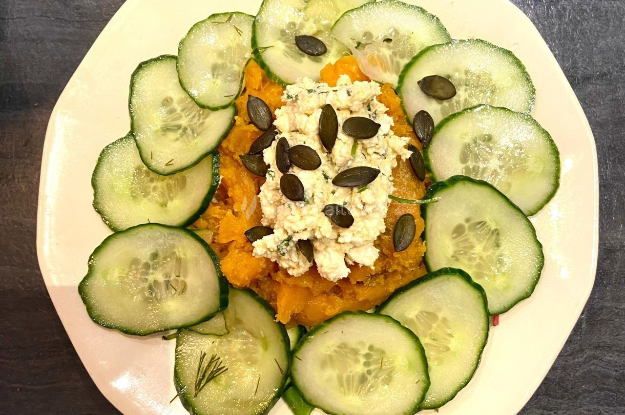 Assiette garnie de purée de légumes orange, fromage émietté, graines de courge et rondelles de concombre disposées en cercle.