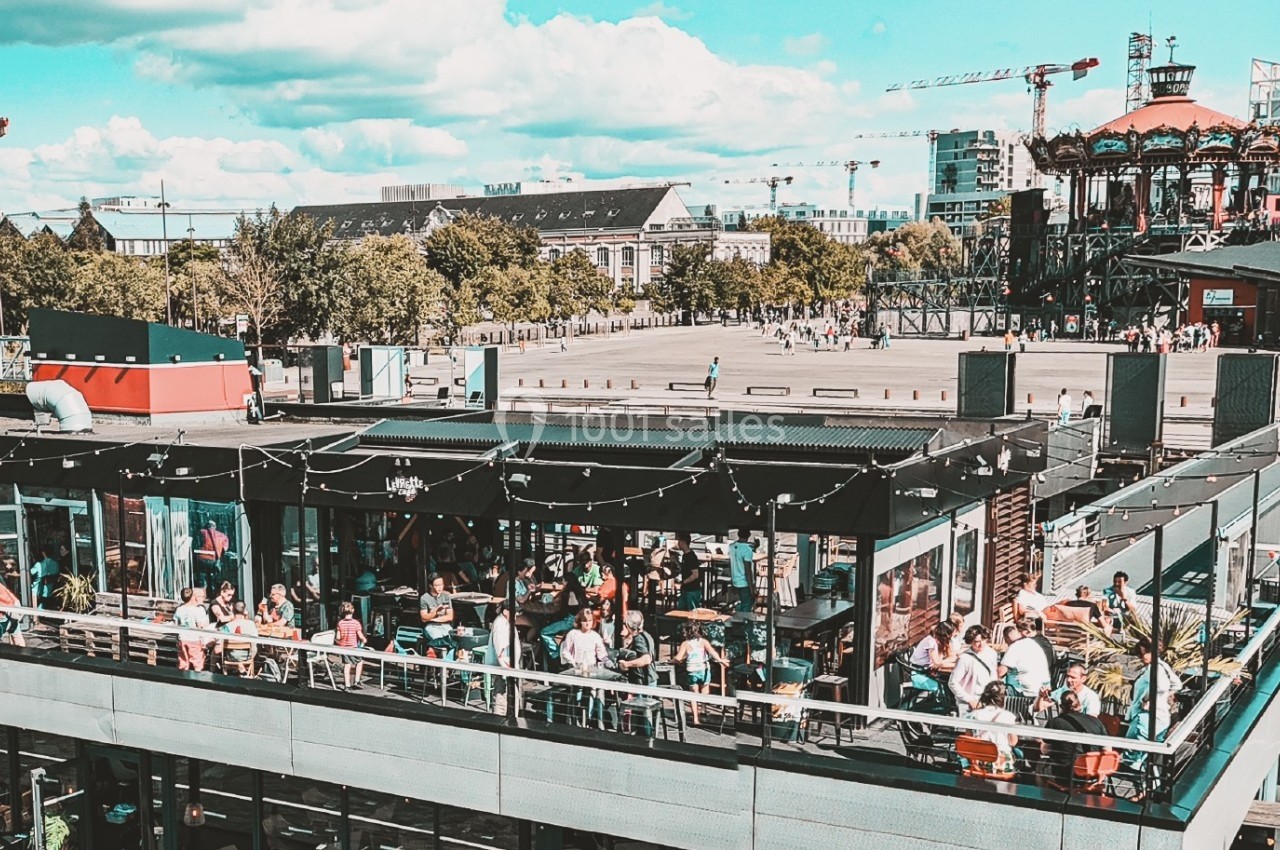 Terrasse animée d'un restaurant avec des clients, située près d'une place et d'un carrousel dans un cadre urbain.