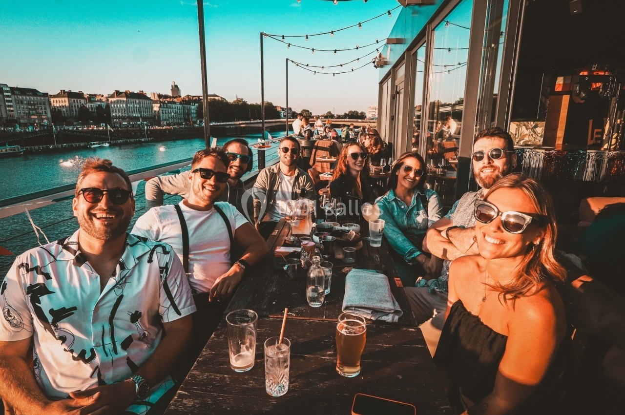 Un groupe de personnes souriantes assises à une table en terrasse au bord d'une rivière par temps ensoleillé.