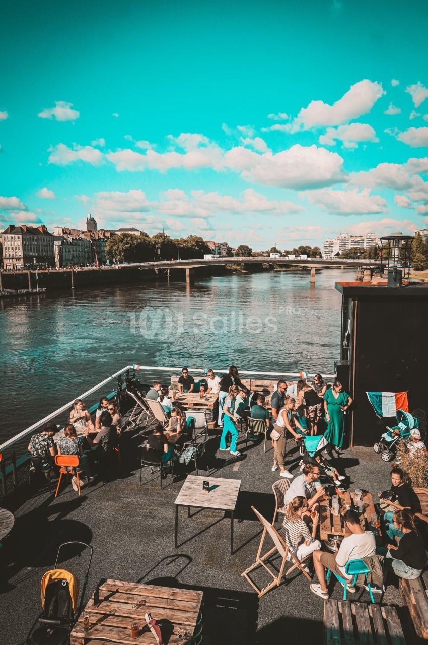 Des personnes assises et debout sur une terrasse au bord d'une rivière, sous un ciel bleu avec quelques nuages.