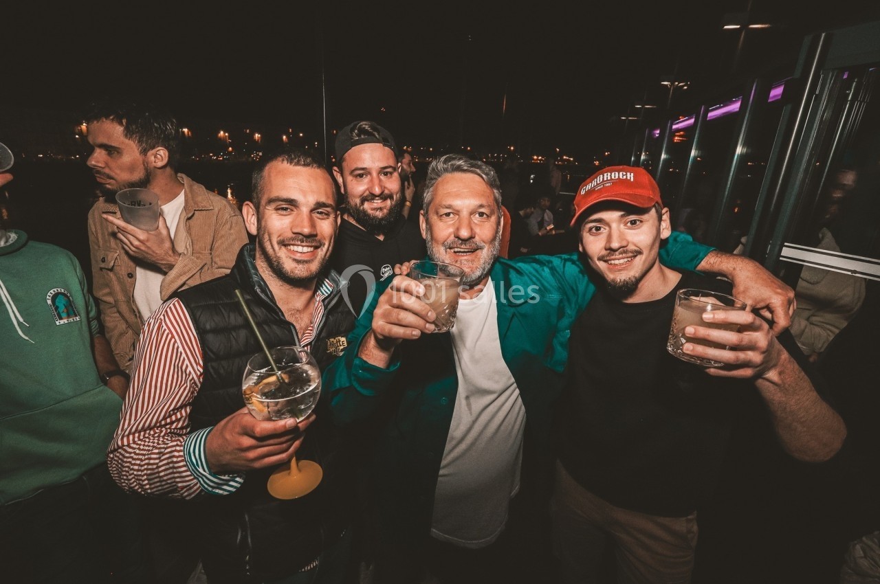 Quatre hommes souriants tenant des verres, posant ensemble lors d'une soirée en extérieur, éclairée par des lumières…