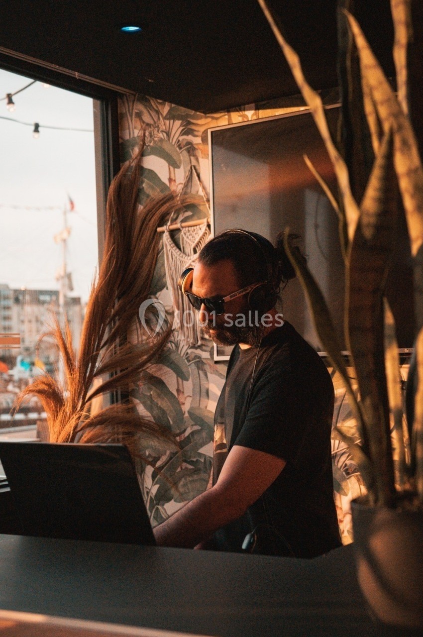 Un DJ portant des lunettes de soleil mixe de la musique dans un espace intérieur avec vue sur un port au coucher du soleil.