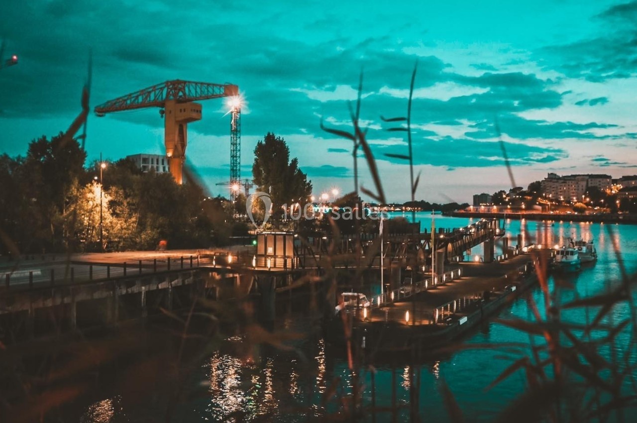 Vue nocturne d'un port avec des lumières reflétées sur l'eau, une grue et des bâtiments en arrière-plan.
