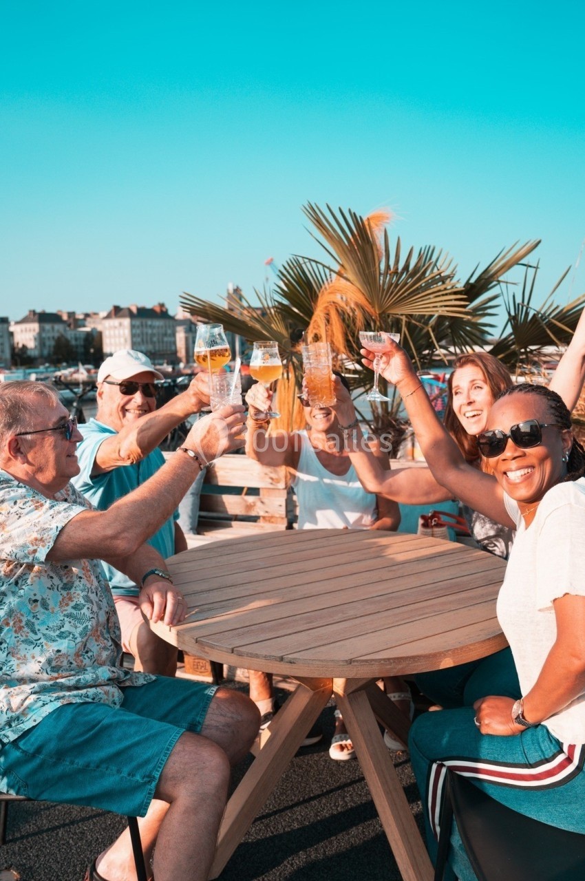 Des personnes souriantes trinquent avec des verres autour d'une table en extérieur, sous un ciel bleu.