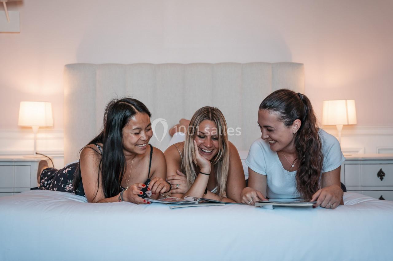 Trois femmes souriantes lisent des magazines allongées sur un lit dans une chambre lumineuse.