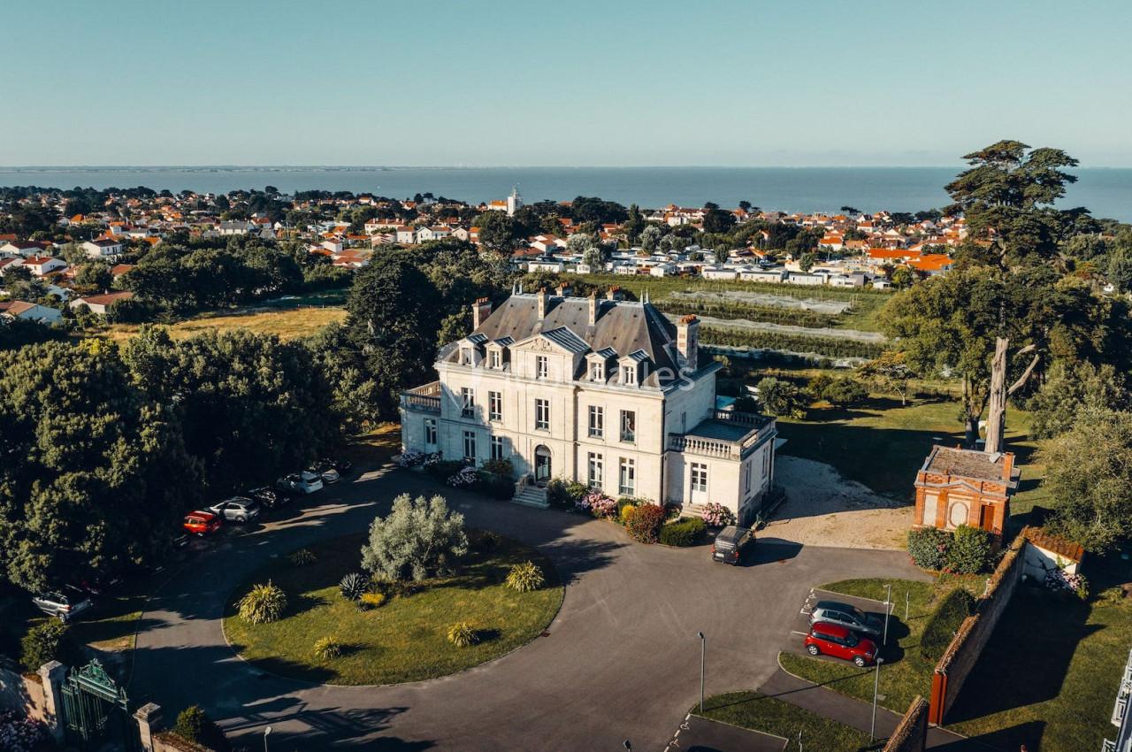 Vue aérienne d'un manoir entouré de jardins, avec une petite ville et la mer en arrière-plan.