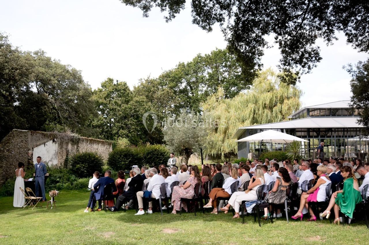 Un groupe de personnes assiste à une cérémonie en plein air dans un jardin, avec des arbres et un bâtiment en arrière-plan.