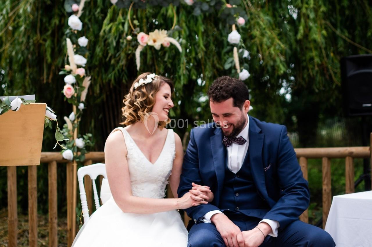 Un couple assis, souriant et se tenant la main lors d'une cérémonie de mariage en extérieur.