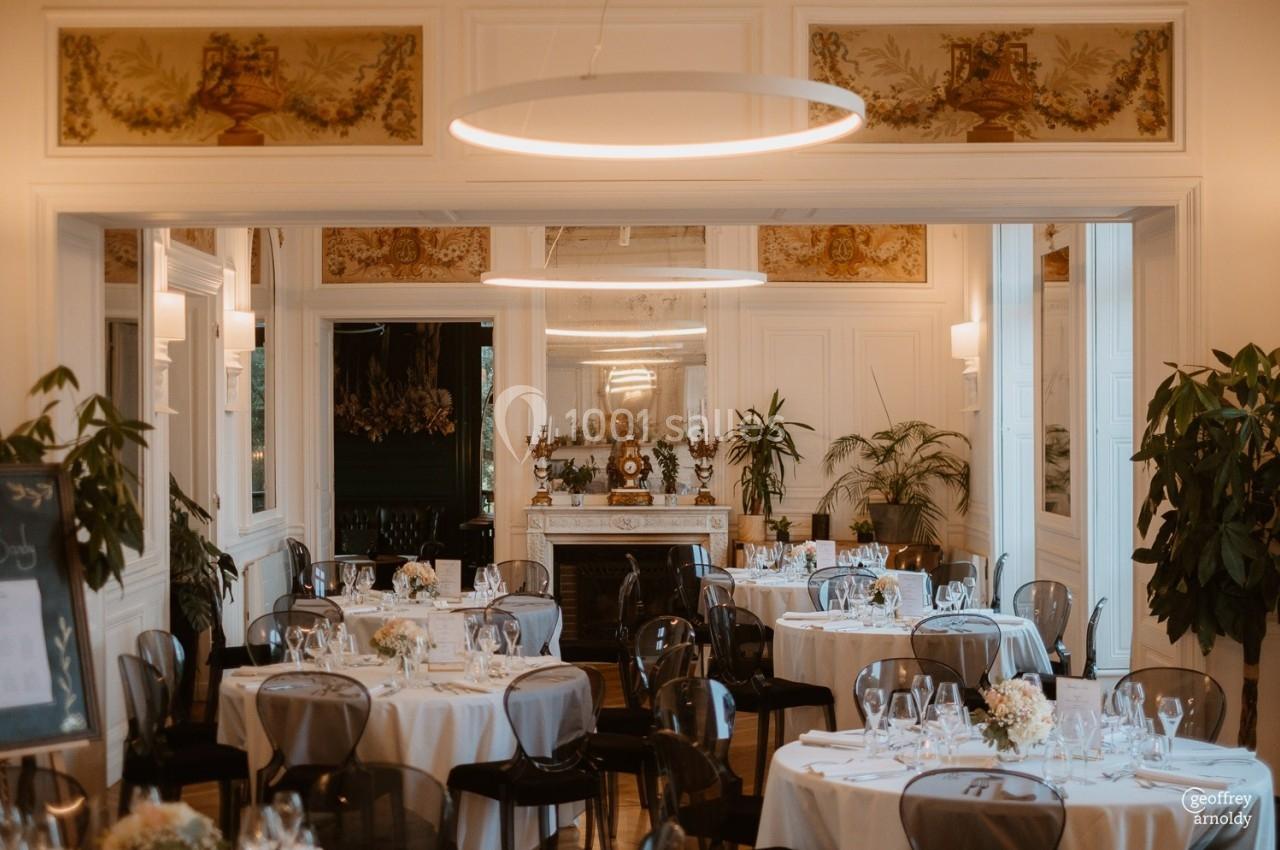 Salle de réception élégante avec tables dressées, chaises transparentes, décorations florales et cheminée en arrière-plan.