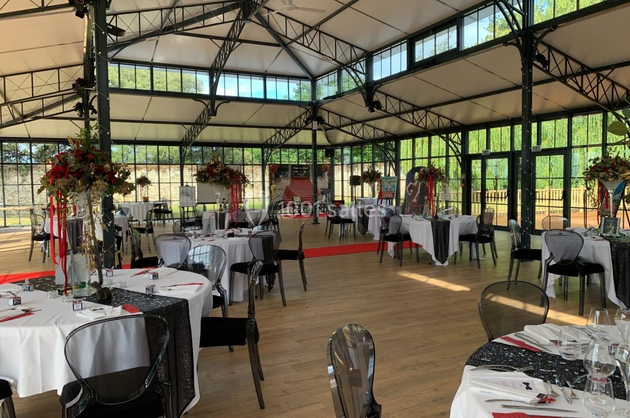 Salle de réception lumineuse avec tables dressées, nappes blanches et décorations florales, entourée de grandes baies…