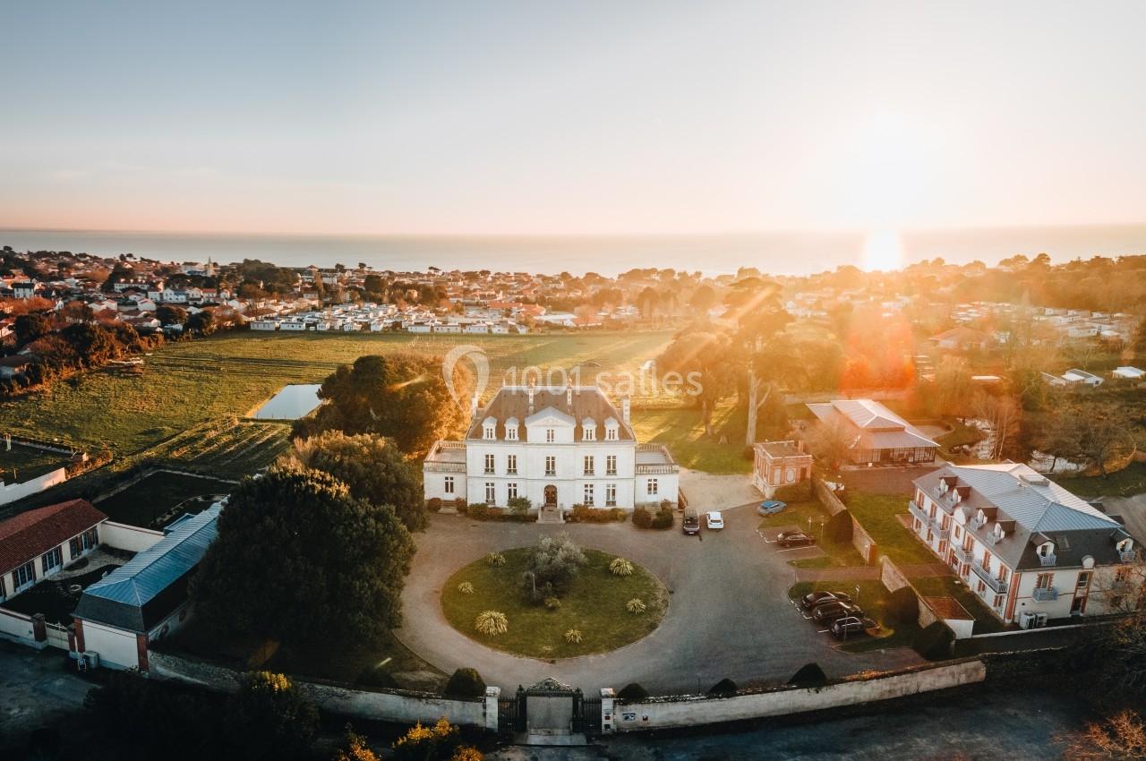 Vue aérienne d'un manoir entouré de jardins, avec un village et la mer en arrière-plan au coucher du soleil.