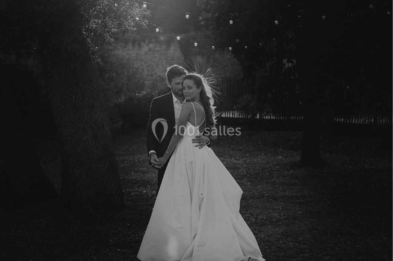 Un couple en tenue de mariage pose dans un jardin sombre, éclairé par des guirlandes lumineuses suspendues.