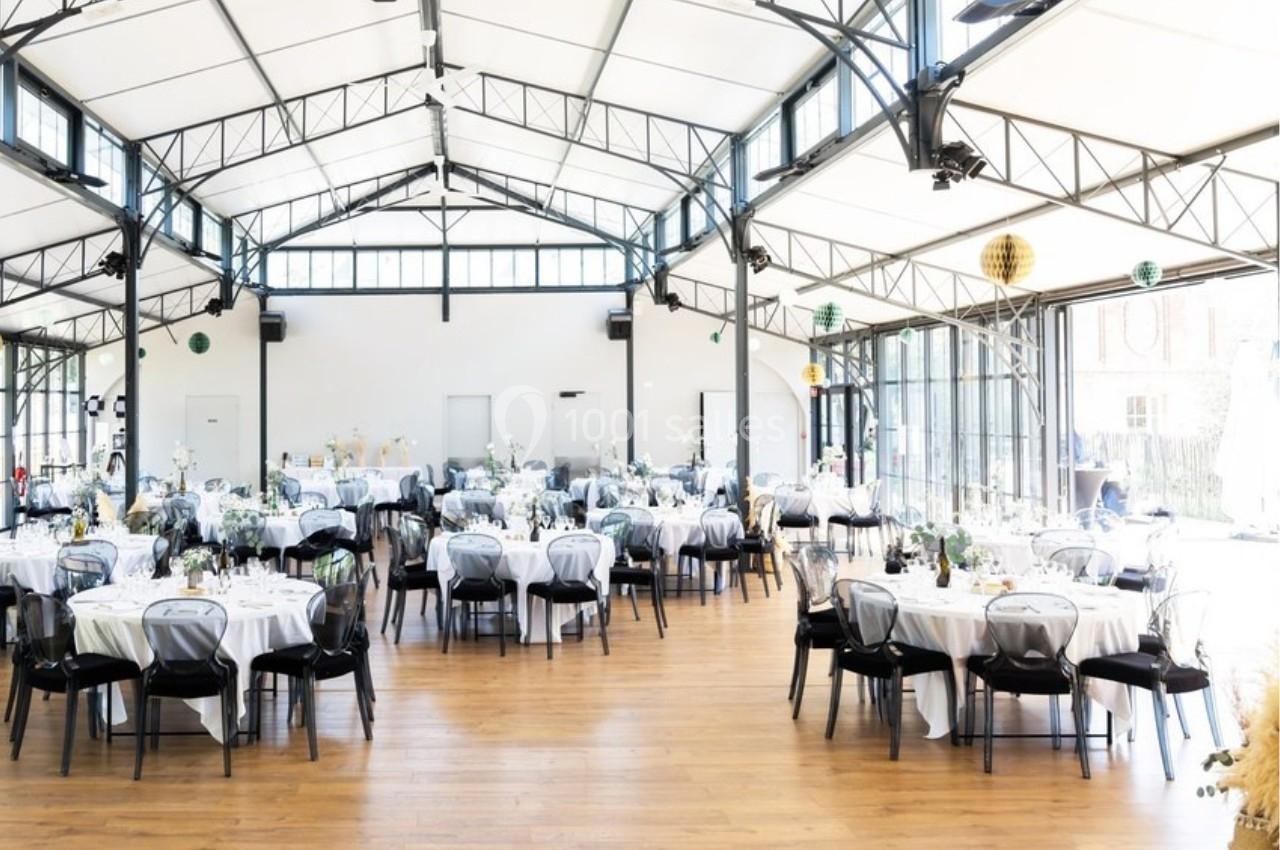 Salle lumineuse avec de grandes baies vitrées, aménagée pour un événement avec des tables rondes dressées.