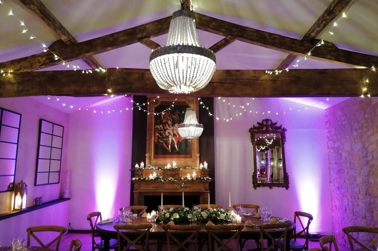 Salle décorée pour un dîner, avec table dressée, guirlandes lumineuses, lustres et éclairage d'ambiance violet.