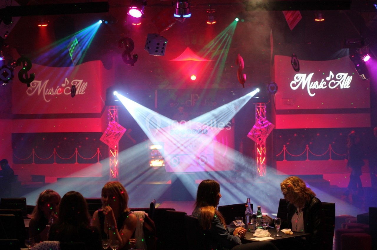 Salle de spectacle avec éclairage coloré, projecteurs, écrans affichant ’Music'All’ et spectateurs attablés au premier plan.