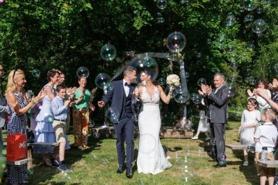 Un couple de mariés souriant marche parmi des invités applaudissant, entourés de bulles de savon dans un jardin verdoyant.
