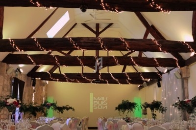 Salle de réception décorée avec des guirlandes lumineuses suspendues, tables dressées et chaises ornées de rubans roses.