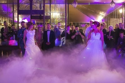 Deux mariées dansent au milieu d'une piste enveloppée de fumée, entourées d'invités dans une salle éclairée.