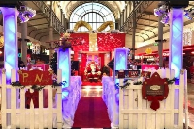 Décor de Noël dans un centre commercial avec Père Noël, sapins, lumières et clôture blanche ornée de décorations.