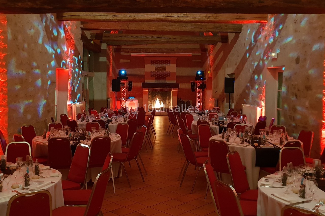 Salle de réception décorée avec des lumières rouges, tables rondes dressées et chaises rouges alignées.