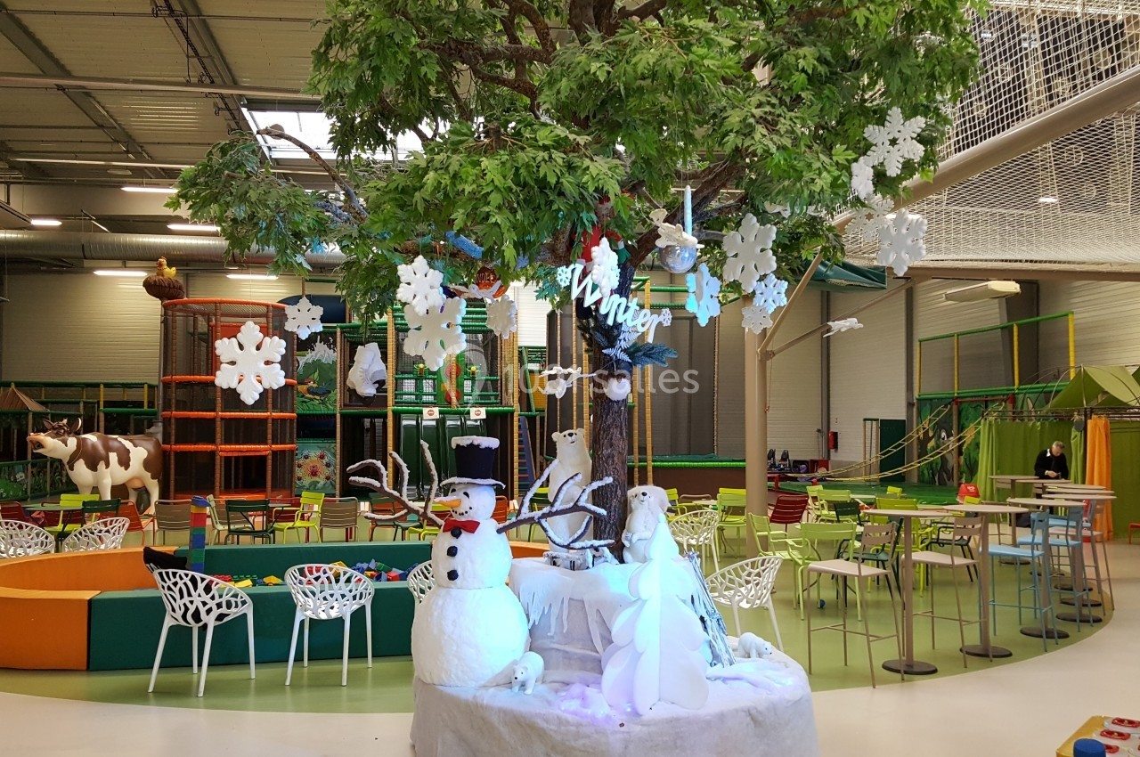 Aire de jeux intérieure avec décorations hivernales, incluant un arbre central, un bonhomme de neige et des tables colorées.