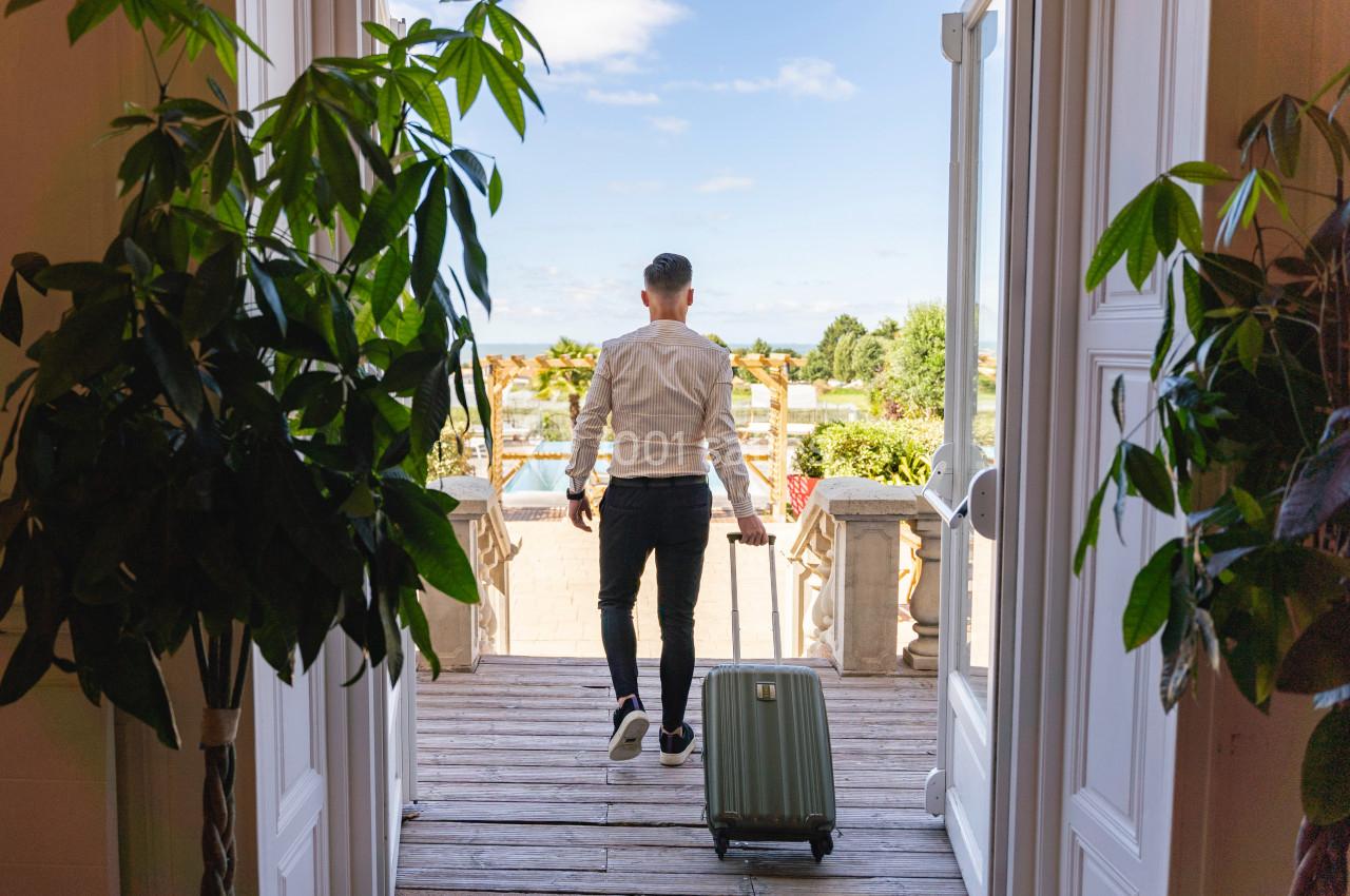 Un homme tirant une valise sort sur une terrasse ensoleillée avec vue sur un jardin et un ciel dégagé.