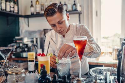 Un homme verse du vin dans une carafe en verre, en extérieur, avec des plantes floues en arrière-plan.