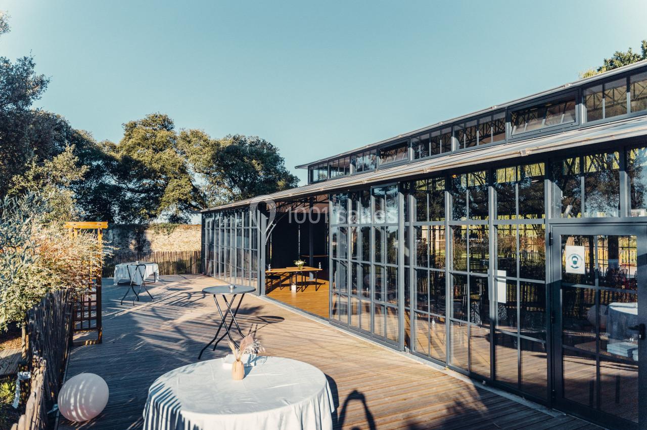 Terrasse en bois avec tables dressées, donnant sur une structure vitrée entourée d'arbres sous un ciel dégagé.