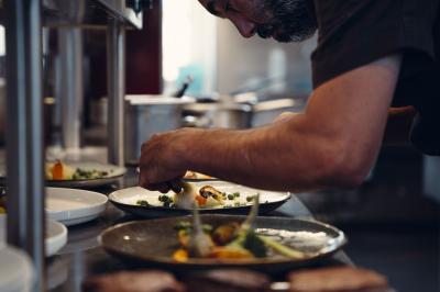 Un homme verse du vin dans une carafe en verre, en extérieur, avec des plantes floues en arrière-plan.