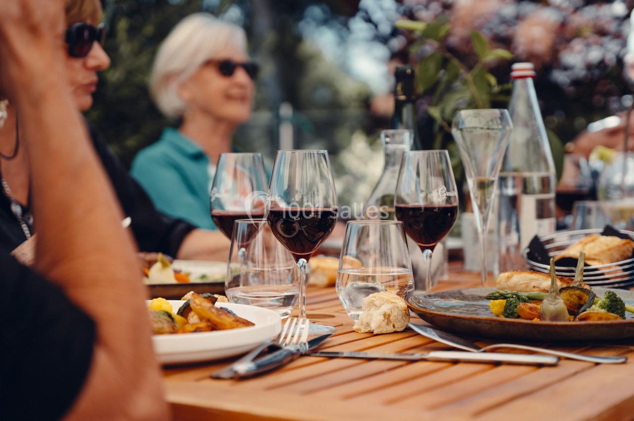 Des convives autour d'une table en extérieur avec des verres de vin, des assiettes et des plats variés.