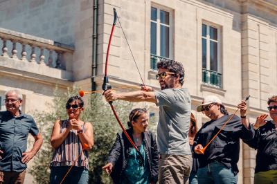 Un homme verse du vin dans une carafe en verre, en extérieur, avec des plantes floues en arrière-plan.