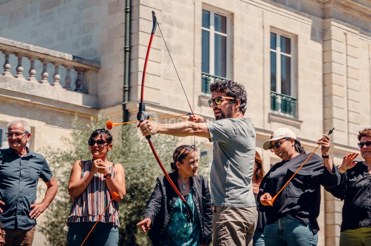 Un homme tire à l'arc en extérieur, entouré de personnes observant devant un bâtiment en pierre.