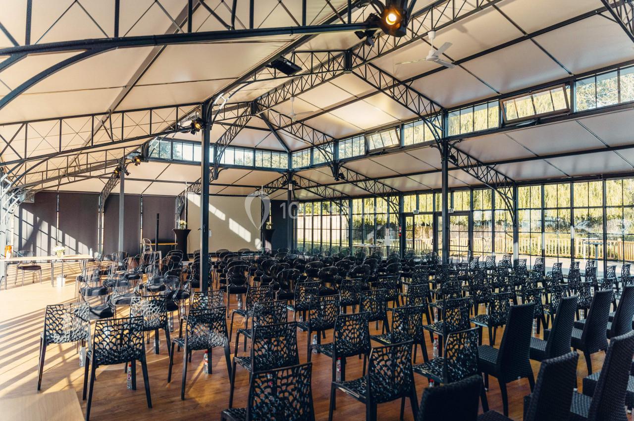 Salle lumineuse avec toit en verrière, rangées de chaises noires disposées face à une scène.