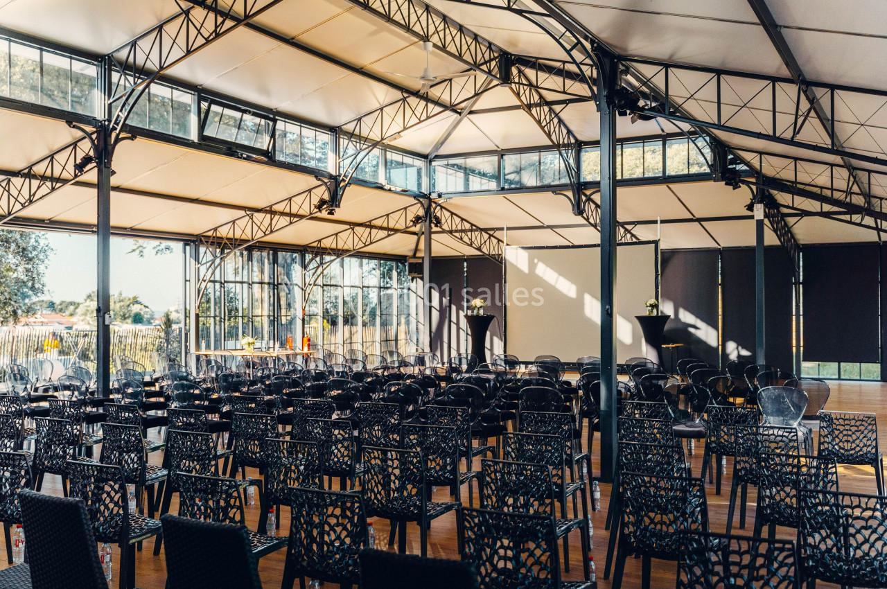 Salle lumineuse avec des chaises noires alignées, un plafond en structure métallique et de grandes baies vitrées.