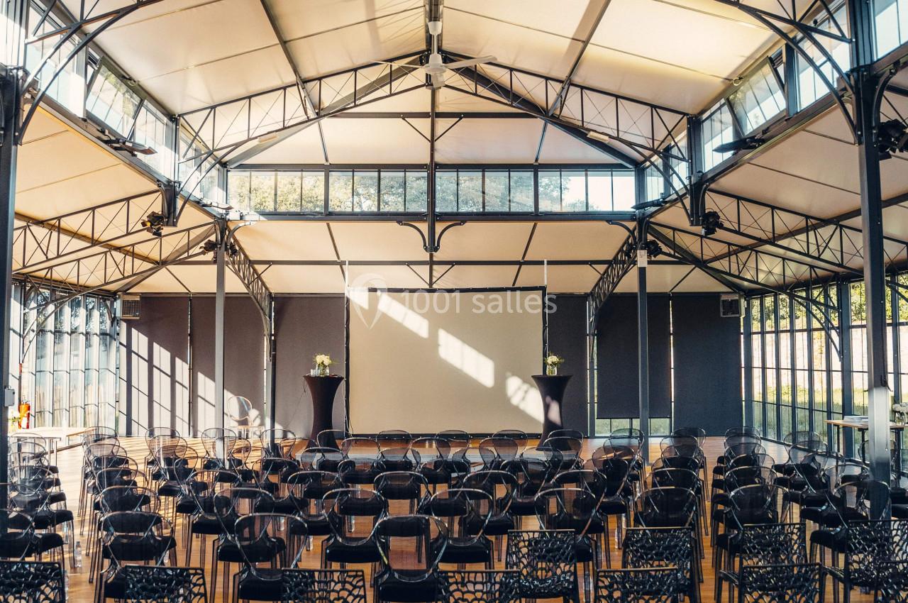 Salle lumineuse avec des rangées de chaises noires devant un écran blanc, sous une structure métallique et un toit en toile.