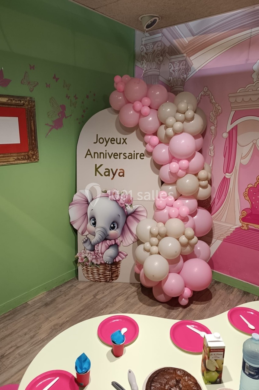 Décoration d'anniversaire avec un éléphant illustré, des ballons roses et beiges, et une table dressée au premier plan.
